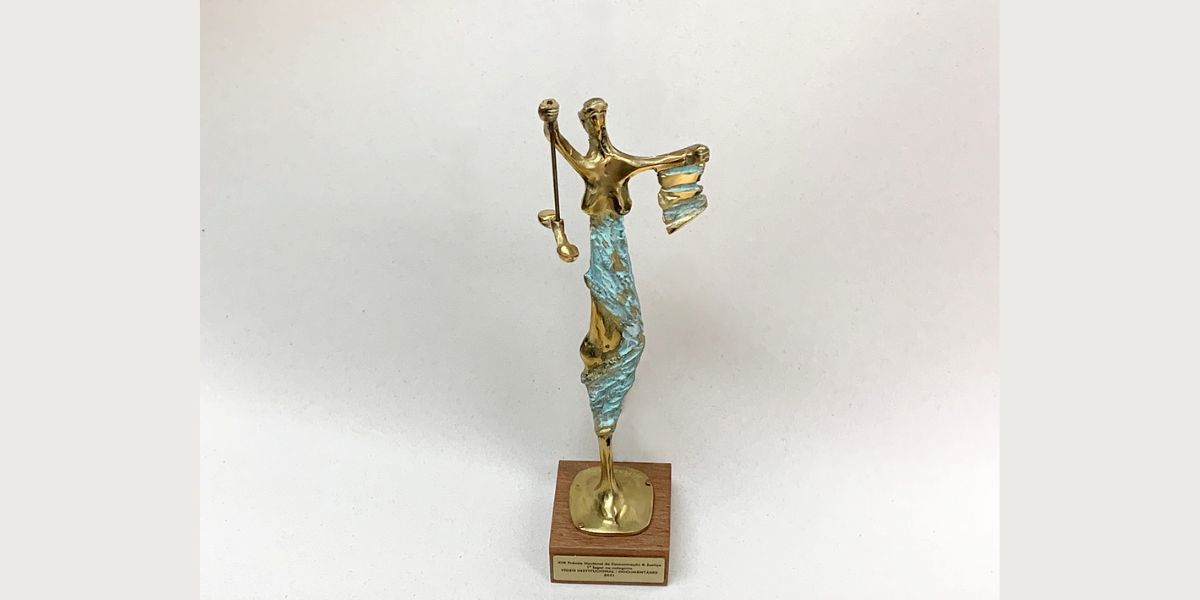 #paracegover - fotografia do troféu do prêmio, que é uma mulher segurando uma balança e um papel, com o cartaz do filme ao fundo.